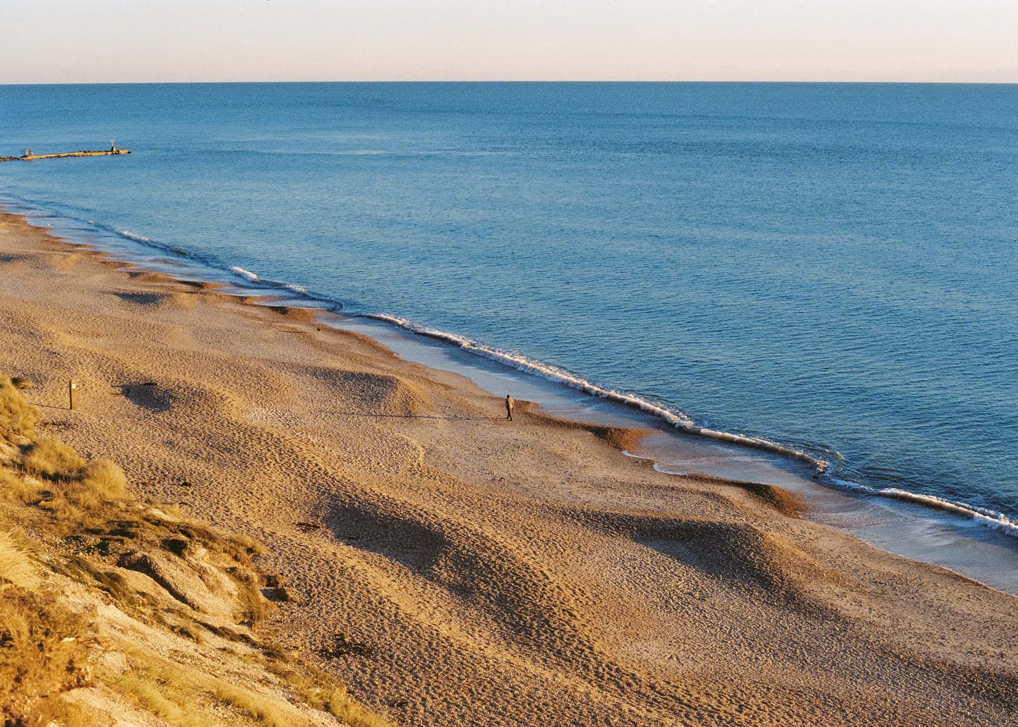 A man walking his dog on the beach at sunset.